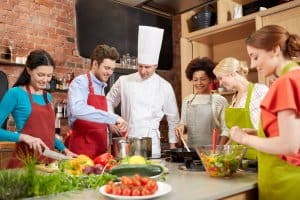 cooking class, culinary, food and people concept - happy group of friends and male chef cook cooking in kitchen